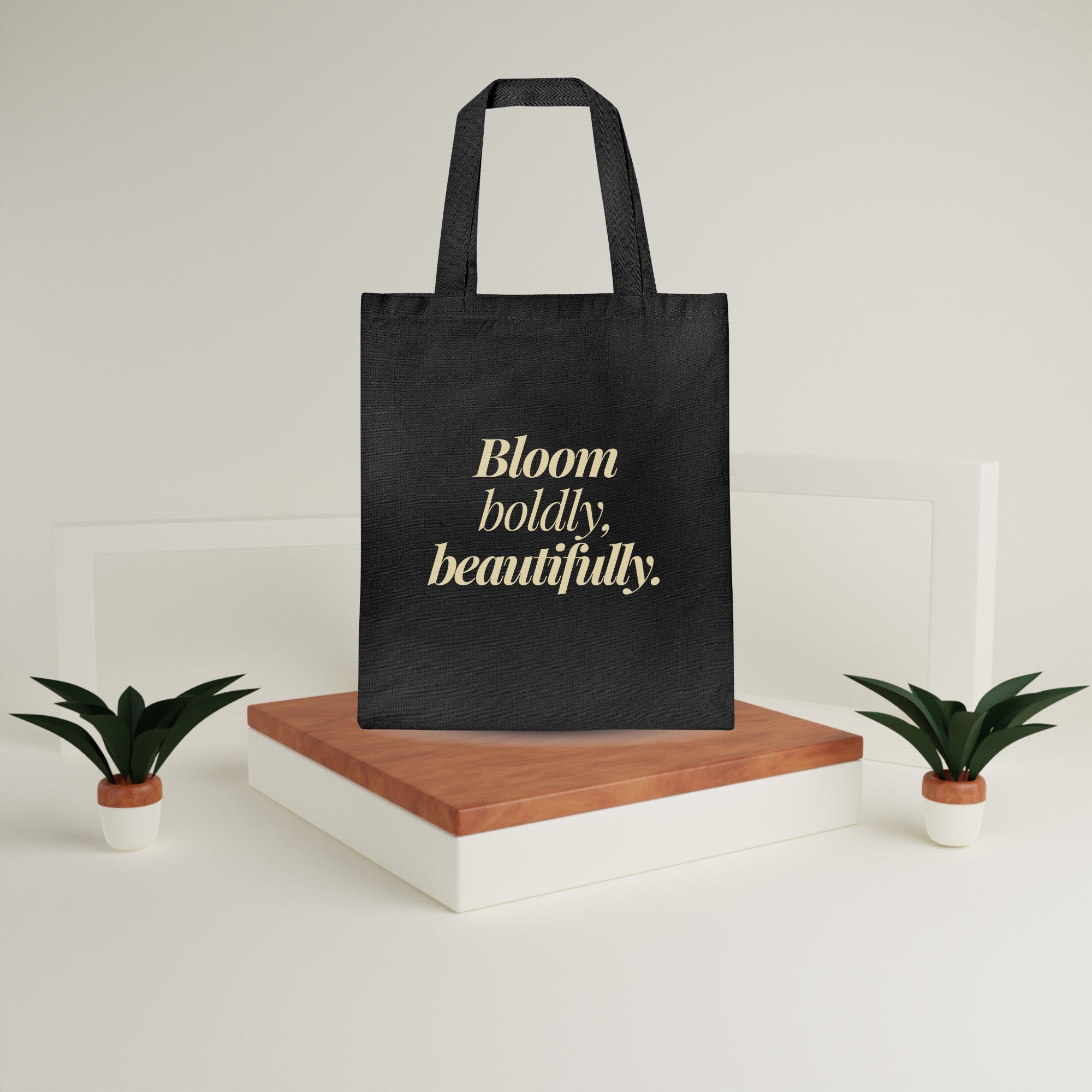 Canvas Tote Bag – Bloom Boldly Beautifully Display Edition Black canvas tote bag with “Bloom boldly, beautifully” in gold text above a wooden platform with plants