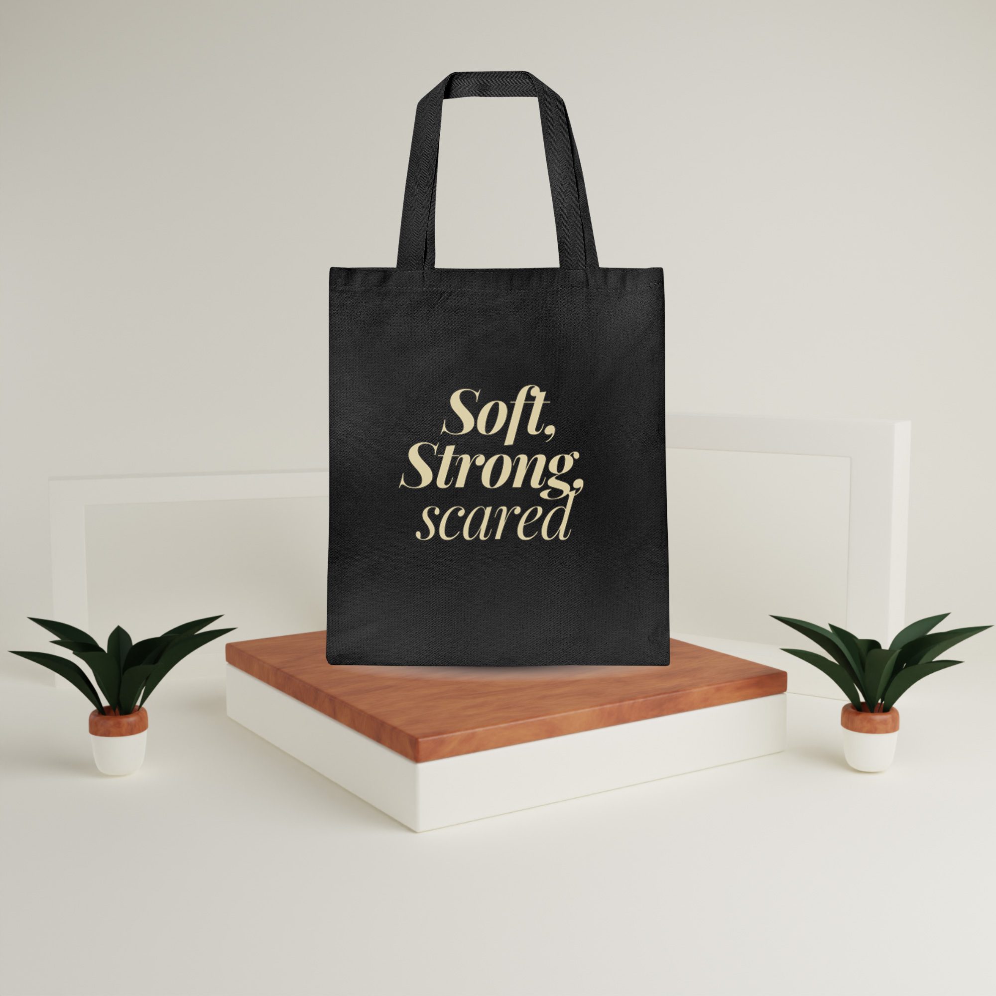 Canvas Tote Bag – Soft Strong Scared Display Edition Black canvas tote bag with “Soft, Strong, scared” in gold text on a wooden platform with plants
