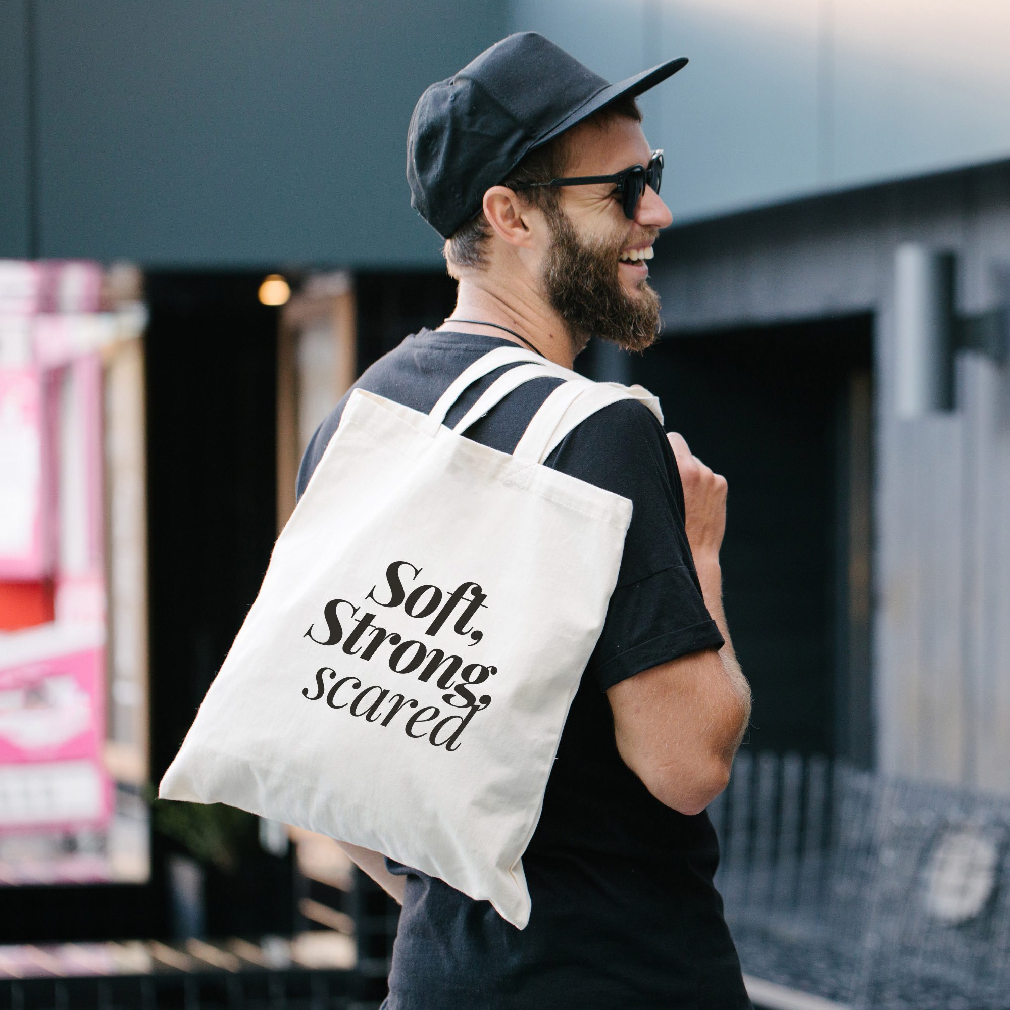 Canvas Tote Bag – Soft Strong Scared Urban Style Person wearing white canvas tote bag with “Soft, Strong scared” in bold black text, standing in front of a modern building