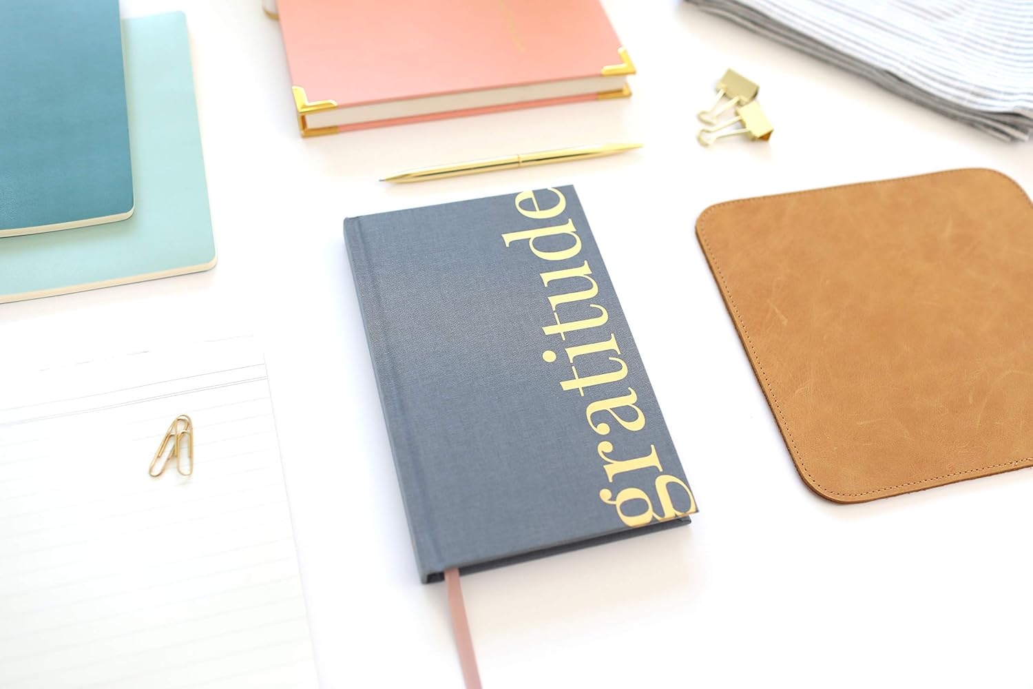 Gratitude Stationery Flat Lay – Organized & Mindful Workspace Flat lay of stationery items including a dark blue “gratitude” notebook, pink and light blue notebooks, gold pen, paper clips, and a striped cloth on a white surface.
