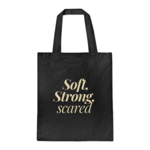 Black canvas tote bag with “Soft, Strong, scared” in beige cursive and serif fonts