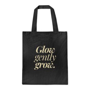 Black canvas tote bag with “Glow gently grow” printed vertically in bold black text