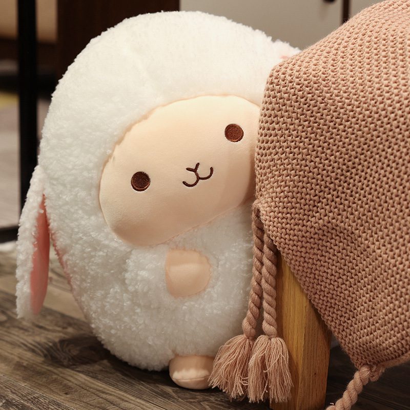 Round Sheep Plushie – Hearthside Hugger Round sheep plushie with peach face and pink ears sitting beside a textured blanket.