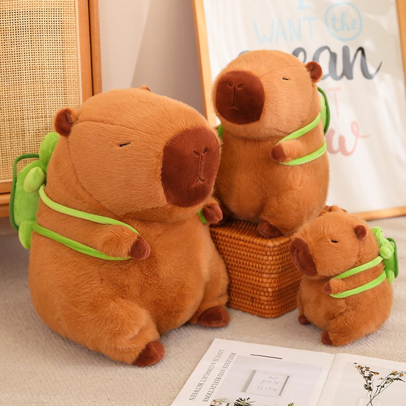 Capybara Plush Toy – Family Pack Edition Three capybara plush toys of varying sizes with green backpacks, arranged in a cozy indoor setting.